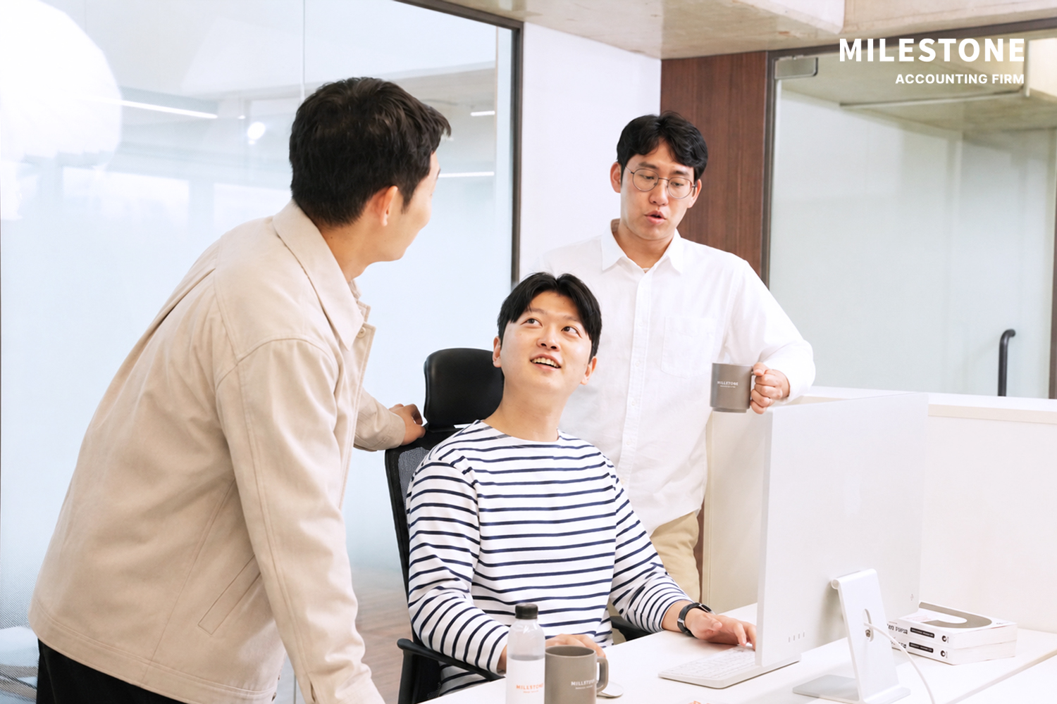 사무실 책상에 앉은 남자와 두 명의 남자가 함께 업무 대화를 나눈다.